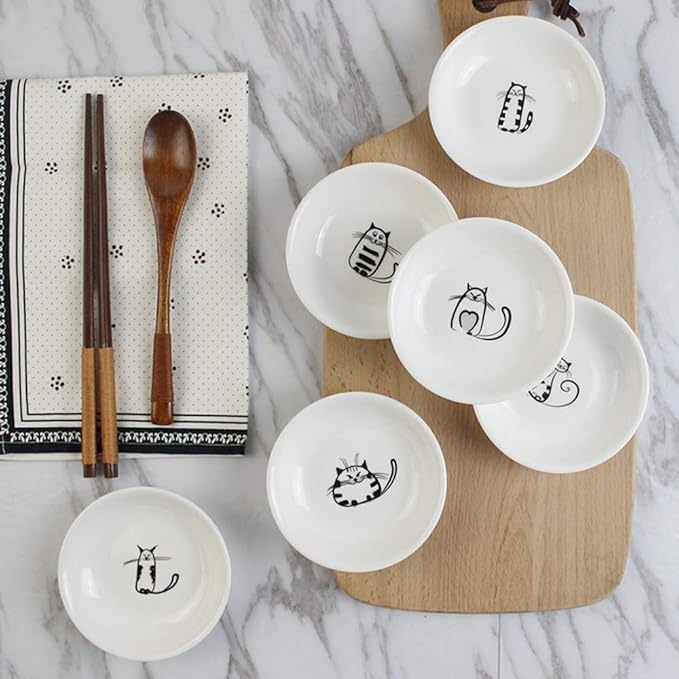 Set of white bowls with cat illustrations on a wooden board, accompanied by chopsticks and a spoon on a marble surface.